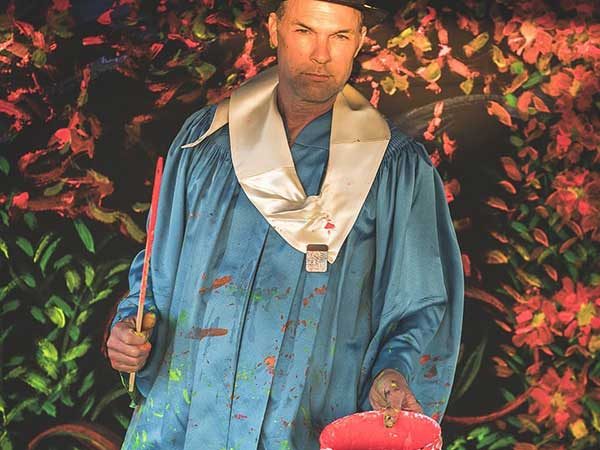 Scott Forrest holding painting bucket and paint stirrer in blue jumpsuit covered in paint.
